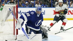 Man wearing hockey goalie uniform and holding a stick blocks a puck trying to go into a net