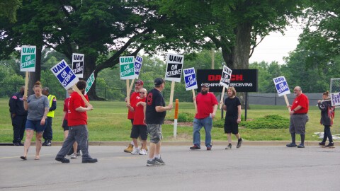 Around ten people stand on the side of the road in front of a chain-link fence and a black sign to the middle right of the image that reads "DAUCH" in red text, below which reads "Shipping & Receiving" in white text with an arrow pointing to the right. The men and women hold different colored signs, but most of them read the same message; "UAW ON STRIKE." One woman's sign to the far right of the image can only be pairly read, it says reads "UNFAIR LABOR PRACTICE" in blue text.
