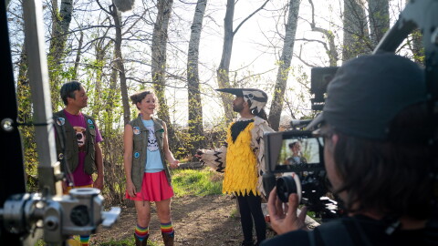 two men and a woman stand in the forest while a TV camera crew films. One of the men is in a yellow and black bird costume