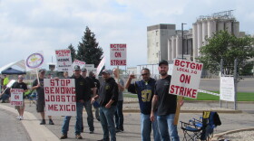 a group of workers stands along a road with the grain silos of the plant in the background. The picketers carry signs with red lettering.