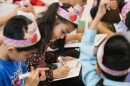 A small girl is in focus among several classmates who are not in focus. She's sitting, wearing a floral patterned shirt and writes with a marker on a portable white board.