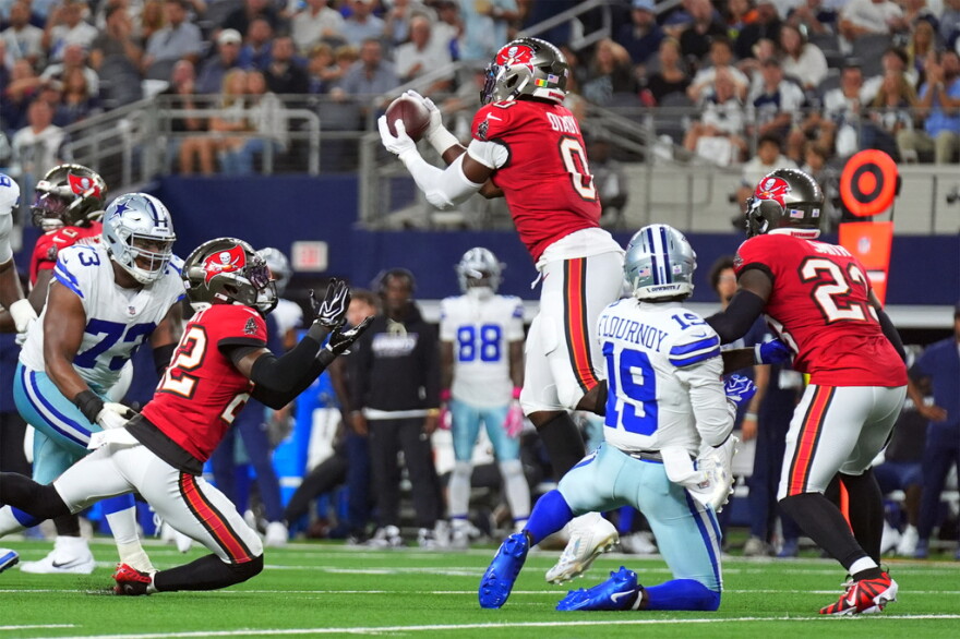 football action with player #0 in red jersey and pewter helmet and white pants jumping to catch the ball. teammates are on each side along with opposing players in white jerseys and gray pants and helmet