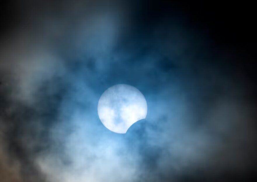 The sun with a portion not visible due to an eclipse. Clouds partially obscure the view. 