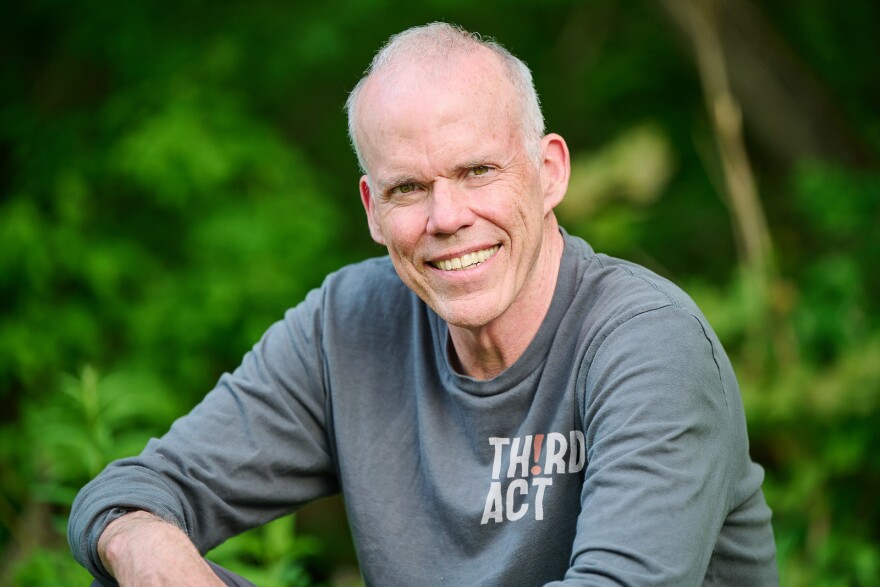 A man sits outside in a gray long-sleeve shirt and smiles at the camera