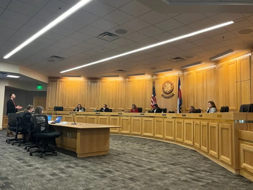Six Weld County Commissioners sit on a dais in a wood-paneled meeting room. A man in a suit addresses the commissioners as two women seated at a desk look on and record what's being discussed.