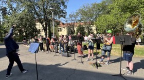 A line of young adults with musical instruments performs outdoors. At the left is a man in dark blue directing the music. 