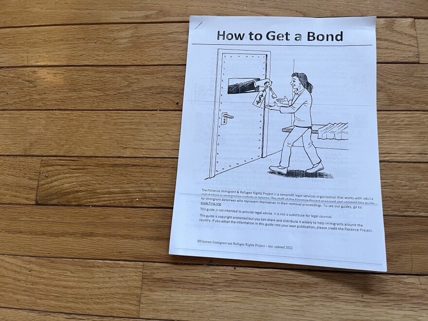 A printed single-page guide titled "How to Get a Bond," photographed lying on a wood floor. The cover features a black-and-white line drawing of a person receiving keys and a money bag through a slot in a heavy, bolted door, suggesting release from detention. Below the illustration, small text identifies the document as produced by the Florence Immigrant & Refugee Rights Project, a nonprofit legal services organization. The text notes the guide is intended for immigrant detainees who represent themselves in removal proceedings and that it is not a substitute for legal counsel.