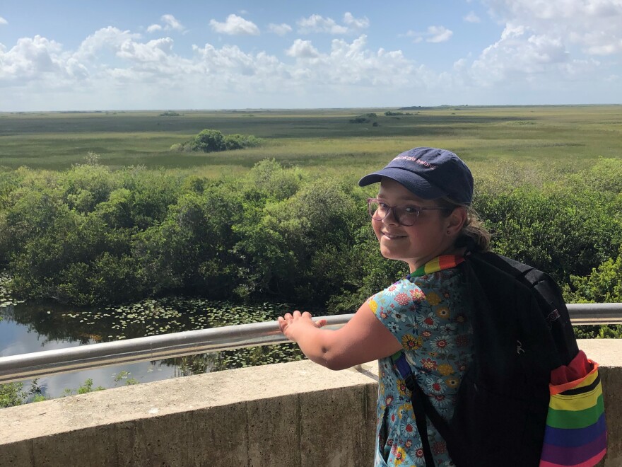 Iris Turmelle wears a flower dress and a black backpack that has rainbow striping on a large front pocket. The straps also rainbow colored. Behind her is a vast grassland and clear blue sky with cumulus clouds.jpg