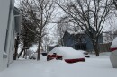 Sidewalks, the road and several cars parked in the street are nearly covered in snow.