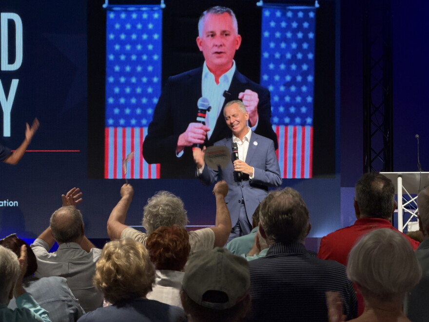A man speaks on stage in front of a large picture of himself, flanked by American flags