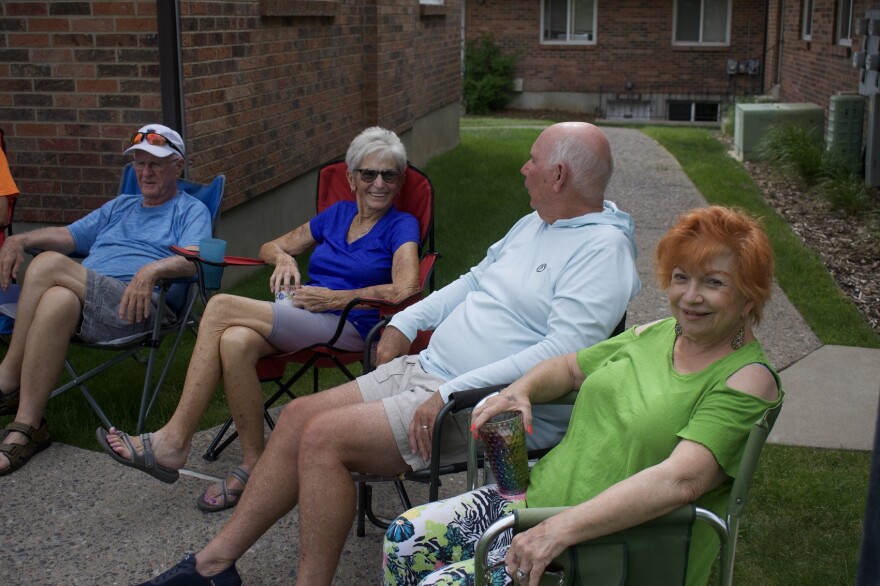 Four senior citizens sit on lawn chairs outside a cluster of small apartment buildings.