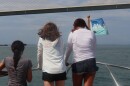 Three pre-teen girls stand on the bow of a boat, holding a metal railing that glints in the sun. They look out onto a body of water and a bridge that rises in front of them. 