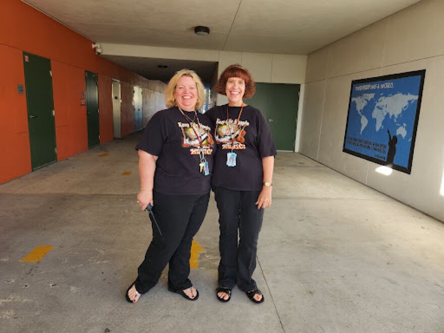 A woman with blonde hair to the left standing arm-in-arm with a woman with brown hair, smiling into the camera and both wearing black t-shirts