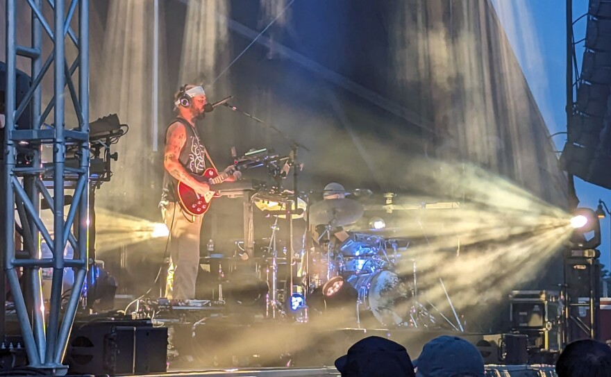 A man plays an electric guitar and sings into a microphone on stage while a colored light shines on him from the front of the stage.