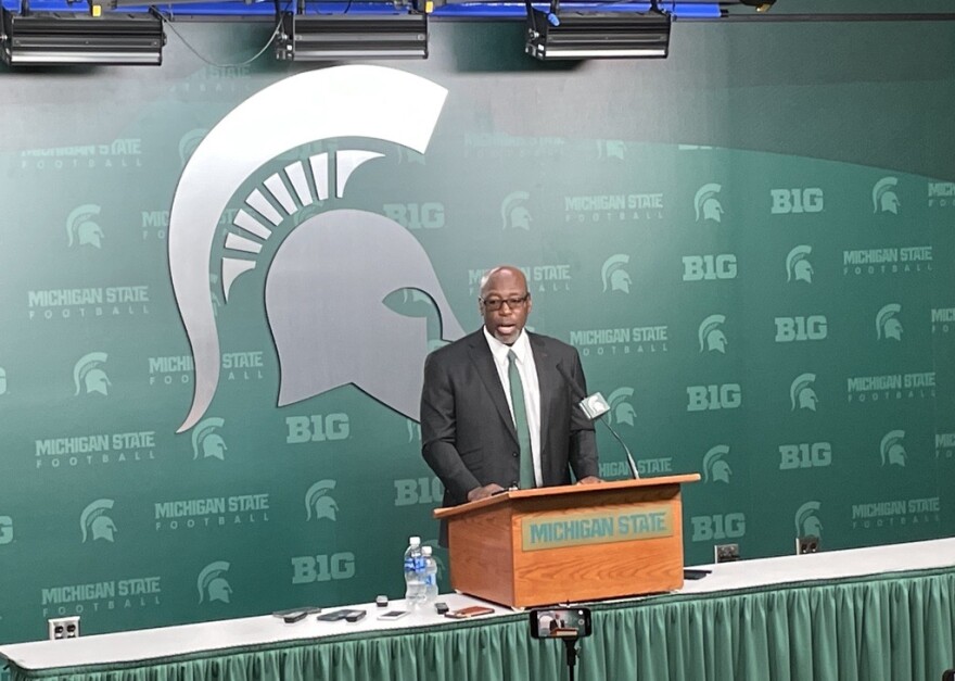 Harlon Barnett, in a suit and green tie, stands in front of the Spartans logo for a press conference.