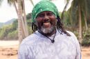 African American man with greyish beard and brown dreads wears a bandana on beach smiles into camera 