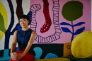 a woman sits with her hands folded on her knees with a tight smile to the camera