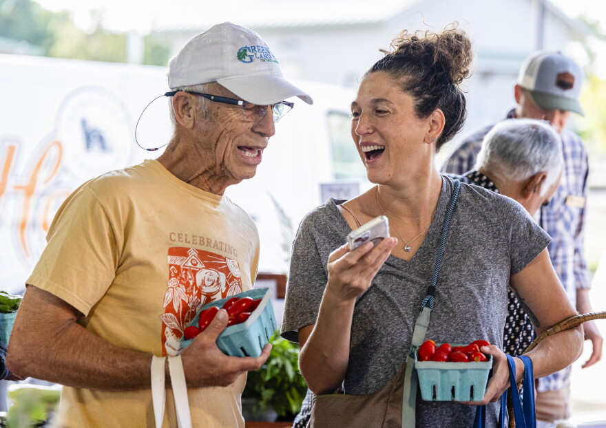 A man wearing a baseball cap and a yellow tshirt stands next to a woman wearing a gray v-neck. She has curly hair in a bun. They are both holding little crates of cherry tomatoes. He leans over to look at her phone. She is smiling and looking at him. 