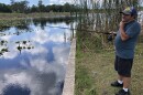 Man standing on grass at the edge of a boat ramp holding a fishing rod with a line going out into the water that is reflecting the blue sky, white clouds, and surrounding trees and plants. He's wearing a cap, t-shirt, shorts and sneakers. 