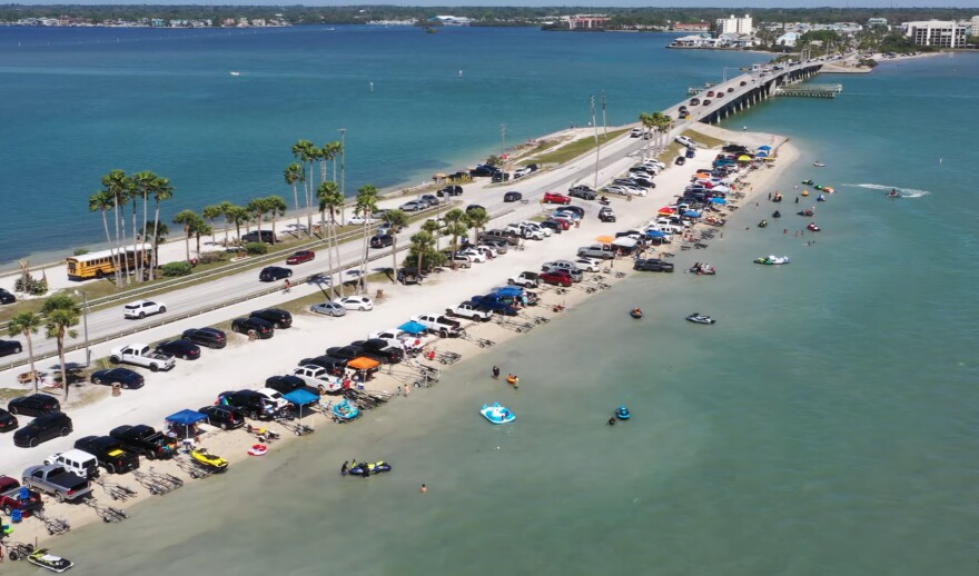 A stretch of road going through a blue body of water. On either side are short sand banks and dozens of parked vehicles and swimmers.