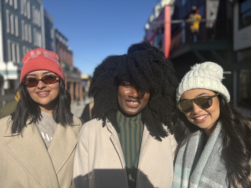 Three women stand outside on a sunny day, wearing coats.