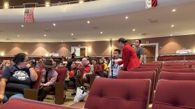 A young boy in a white t-shirt speaks into a microphone held by a woman in a red suit jacket in an auditorium with people seated nearby.