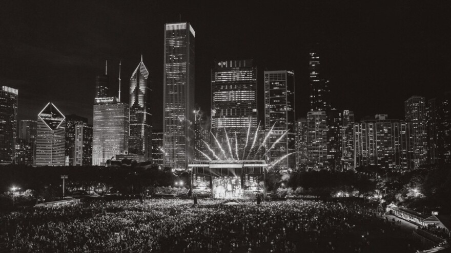 A black-and-white photo of a large park filled with people watching a brightly lit stage at night with a city skyline behind the stage.