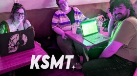 Three people in a coffee shop smiling at the camera. There's an artificial, purplish hue to the picture, and the letters KMST in the bottom foreground.