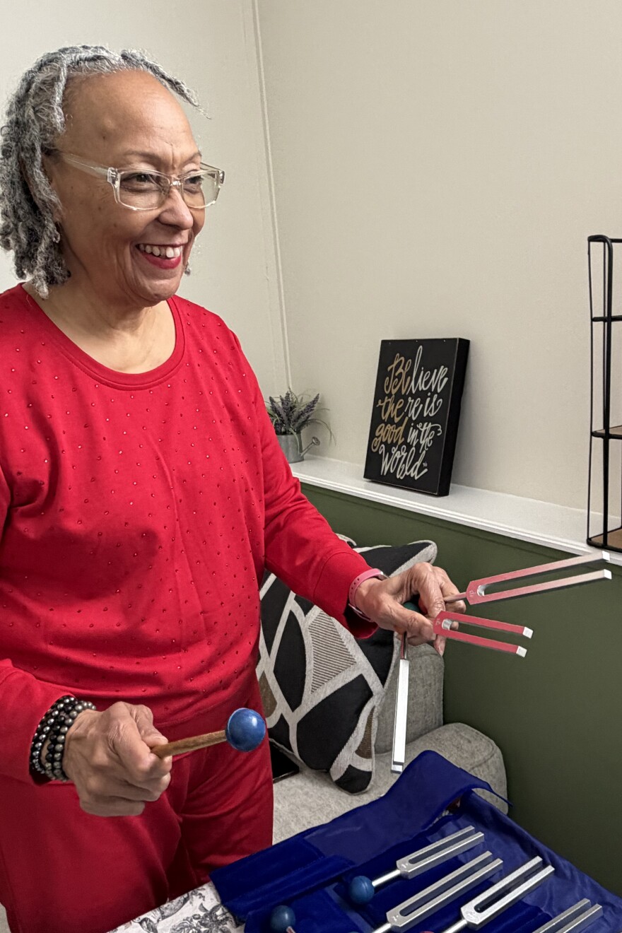 Philana Crite wears a read long sleeve shirt and red pants with beaded bracelets on her right wrist and a watch on her left. She stands to the left side of the image, smiling at a three-quarter angle from the camera. She holds two tuning forks in her left hand and a mallet in the right as she demonstrates their use in spiritual healing. On the table in front of her is a blue velvet carrying case with more tuning forks inside. Behind her is a gray chair with an abstract patterned throw pillow.