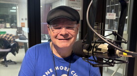 A man sitting and facing forward and smiling near a microphone, wearing a hat and a blue shirt