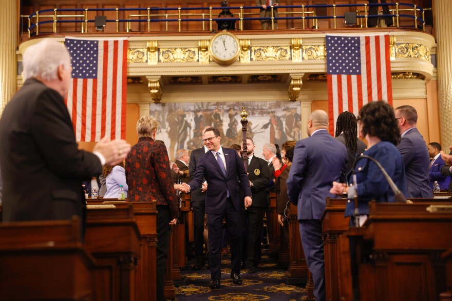 A man in a suit smiles and shakes hands as he walks down an aisle between desks in the Pennsylvania House chamber as legislators stand and applaud.