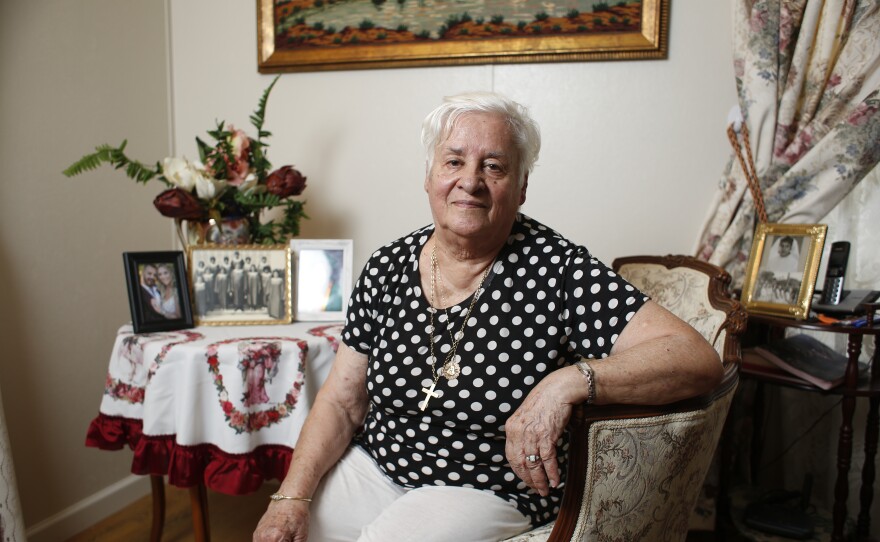 An elderly woman with polka dot shirt sitting in a chair with pictures behind her 