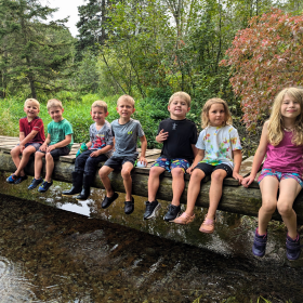 Seven young students sit on a bridge over a wide creek with their feet dangling over the edge.