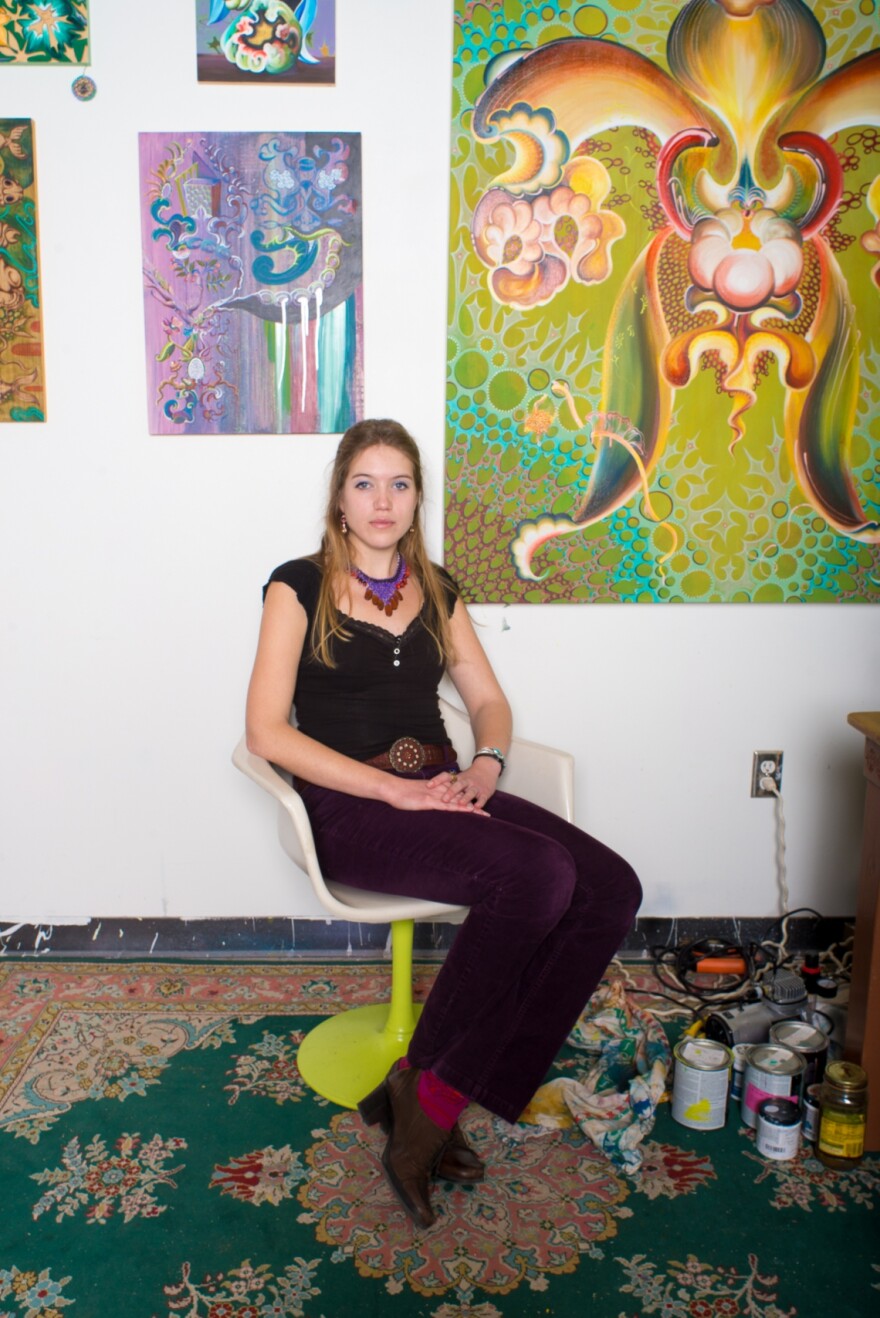 A person seated in front of paintings hanging on a white wall, paints in can on the floor