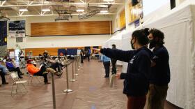 Two officials point while dozens of people sist in foldout chairs