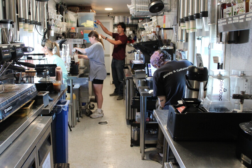 A man reaches for something up high while a woman takes a coffee order through a drive-thru window on the left-hand side. On the right-hand side, a man stands ready to take another order. A fourth employee prepares drinks. 
