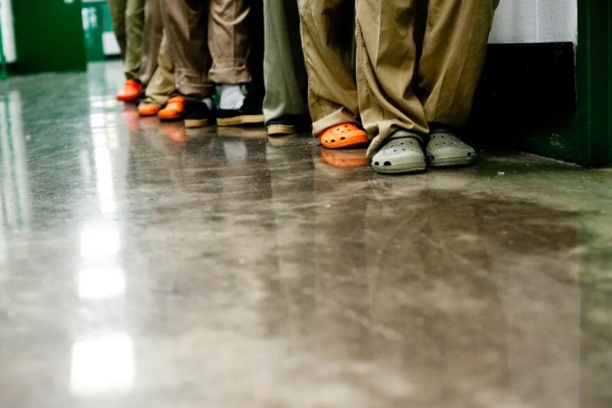 Prison inmates stand lined up, but are seen only from the knee down. Most are wearing Croc-type shoes and baggy pants.