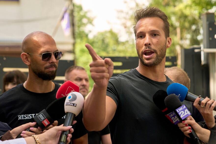 A blad white man wearing sunglasses on the left as his brother, a white man with a receding hairline points a finger outward as he’s surrounded by news microphones.