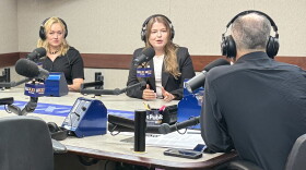 Three people wearing headphones sit at a table in a radio talk studio: a woman at left has long blonde hair and is wearing a short-sleeved black shirt and beaded necklace; a woman at center has long brown hair and is wearing a navy blazer over a white top; a man with very short dark hair at right is sitting with his back to the camera and wearing a navy shirt.