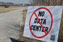 Exterior daytime image of a white sign stuck in the ground. It has a large red circle with a slash over the words "No Data Center." A highway can be seen in background where some cars are moving away.