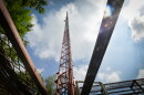 A tall, skinny steel radio tower, painted red and white is seen from below against a cloudy, blue sky. Metal framework near the base is in the foreground.
