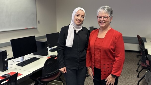 Two women standing in a classroom between rows of students desks and computers. The women on the left is wearing a dark suit with white hijab, the woman on the left is wearing a red top and jacket with blank pants.