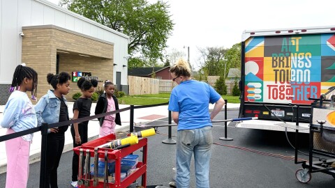  A group of kids gather in front of Kacey McCreery’s glassblowing demo. She’s standing in front of Toledo’s YMCA, holding a rod with molten glass, the furnace burning bright in the right corner.