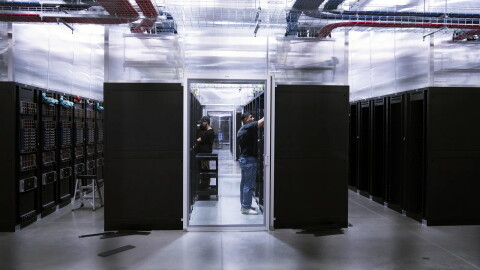 Two people work inside a glass-walled server room surrounded by tall black server racks in a bright, modern data center with overhead cabling—an environment where sustainability is prioritized through the use of hydropower to efficiently run the facility.