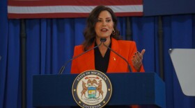 Whitmer, wearing an orange blazer over a dark shirt, stands and talks at a lectern with the state seal on it. There is a blue curtain in the background