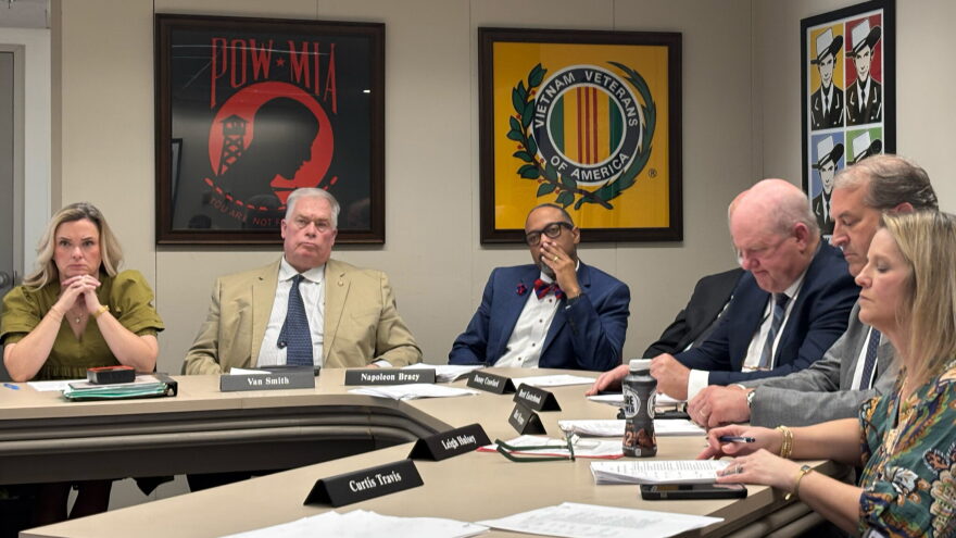 Seven Alabama lawmakers sit around a table. Military-themed posters hang on the wall behind them.