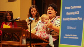 A woman with a baby in a front carrier speaks passionately at a podium in a church while two other women look on. A blurred sign in the foreground reads," Press conference opposing CCS Facility closure plan" with smaller, unreadable print below. 