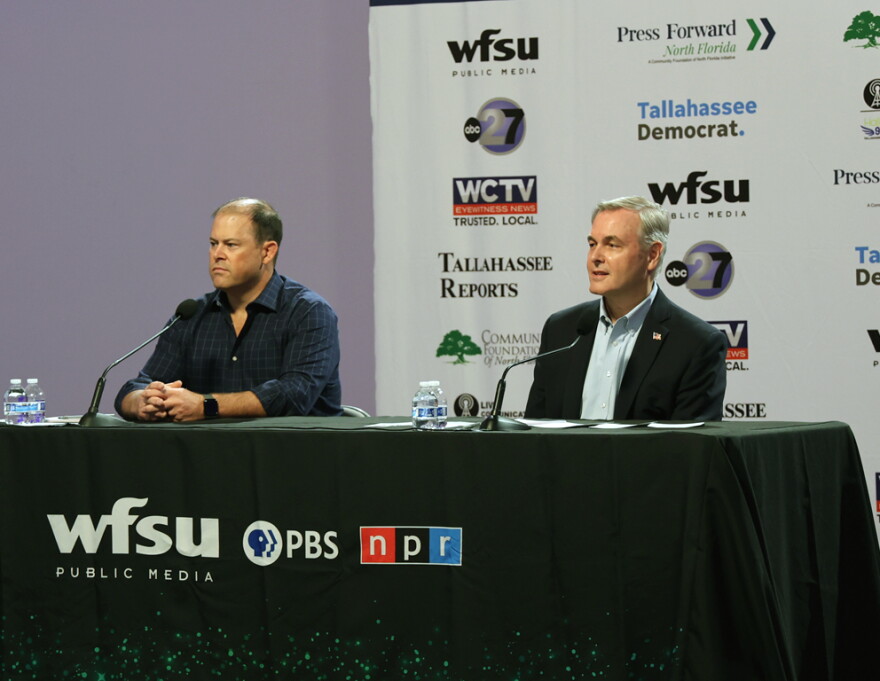 Two men sit at a table in front of microphones staring forward.