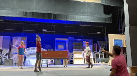 Three children are standing on a stage while the director is guiding their movements from the audience section.