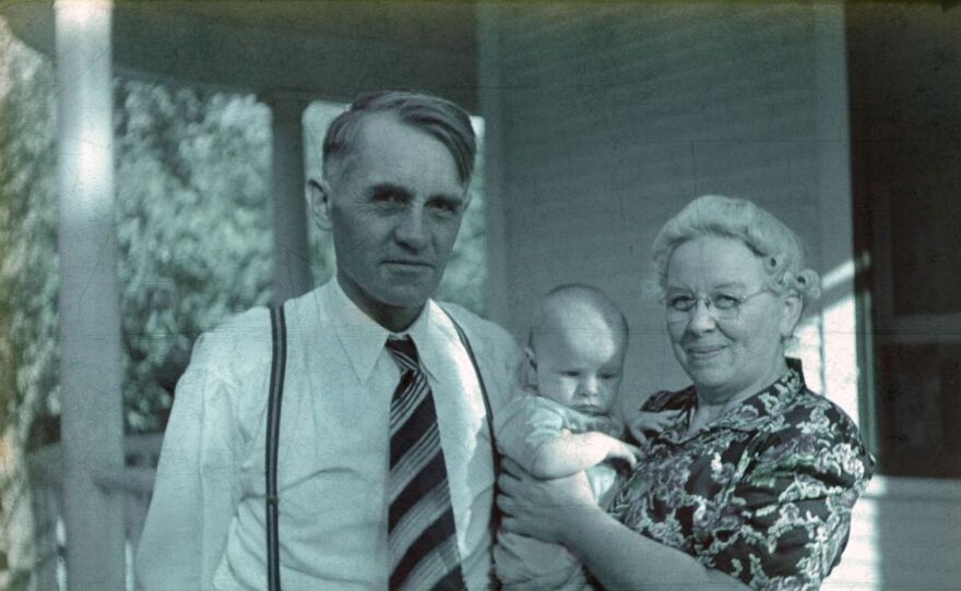 A black and white vintage photo shows a dressed up older man and woman holding a baby.